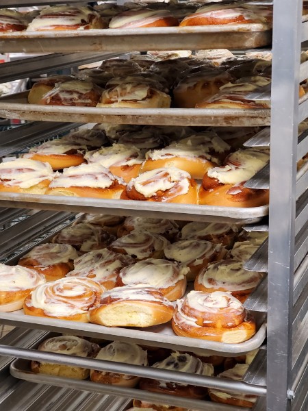 Sheet pans of iced cinnamon rolls stacked down a bakery rack, still glossy from the oven.